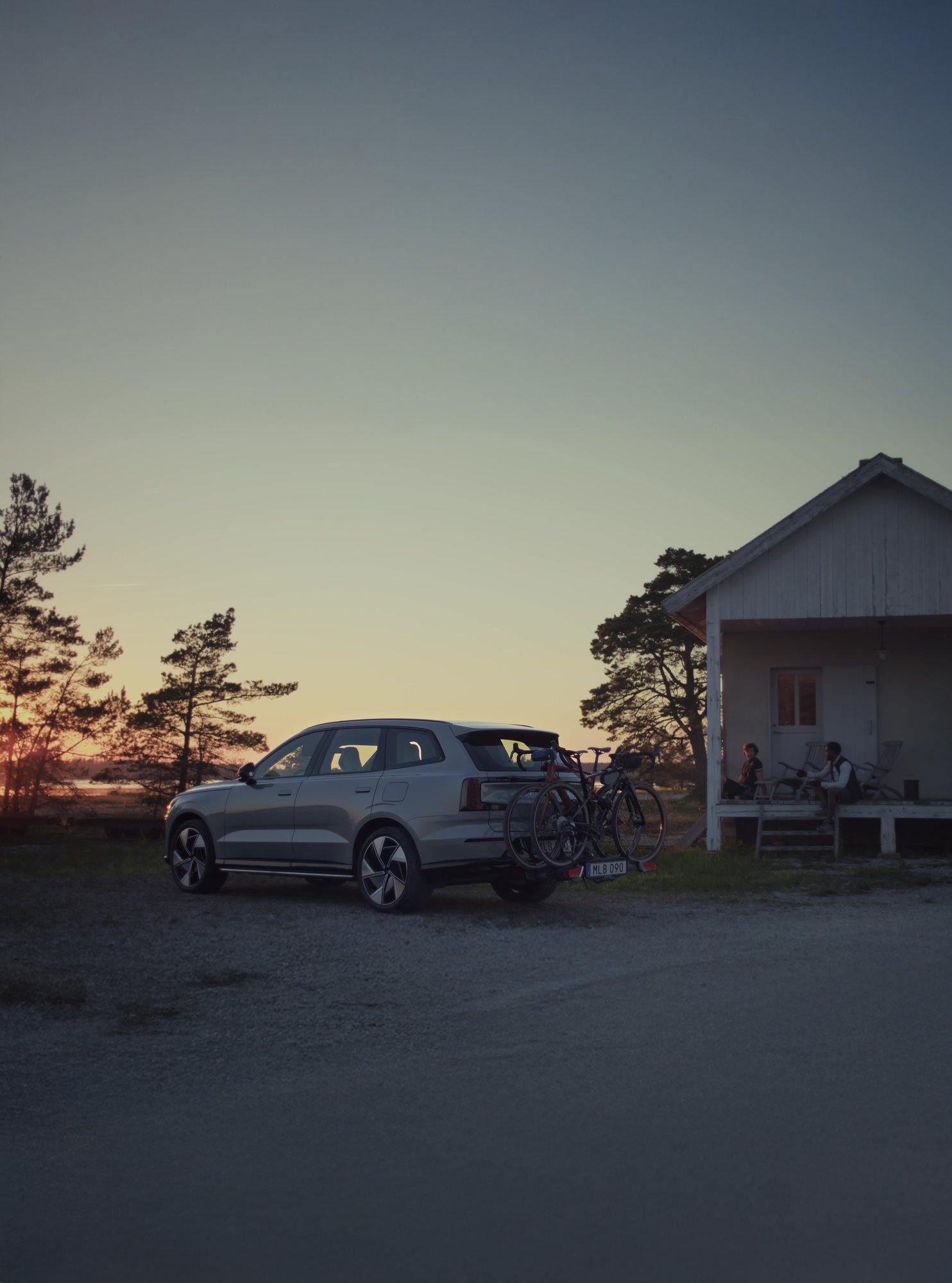 Volvo EX90 sett skrått bakfra i solnedgang, med sykkelstativ montert på hengerfeste og hytte og solnedgang i bakgrunnen