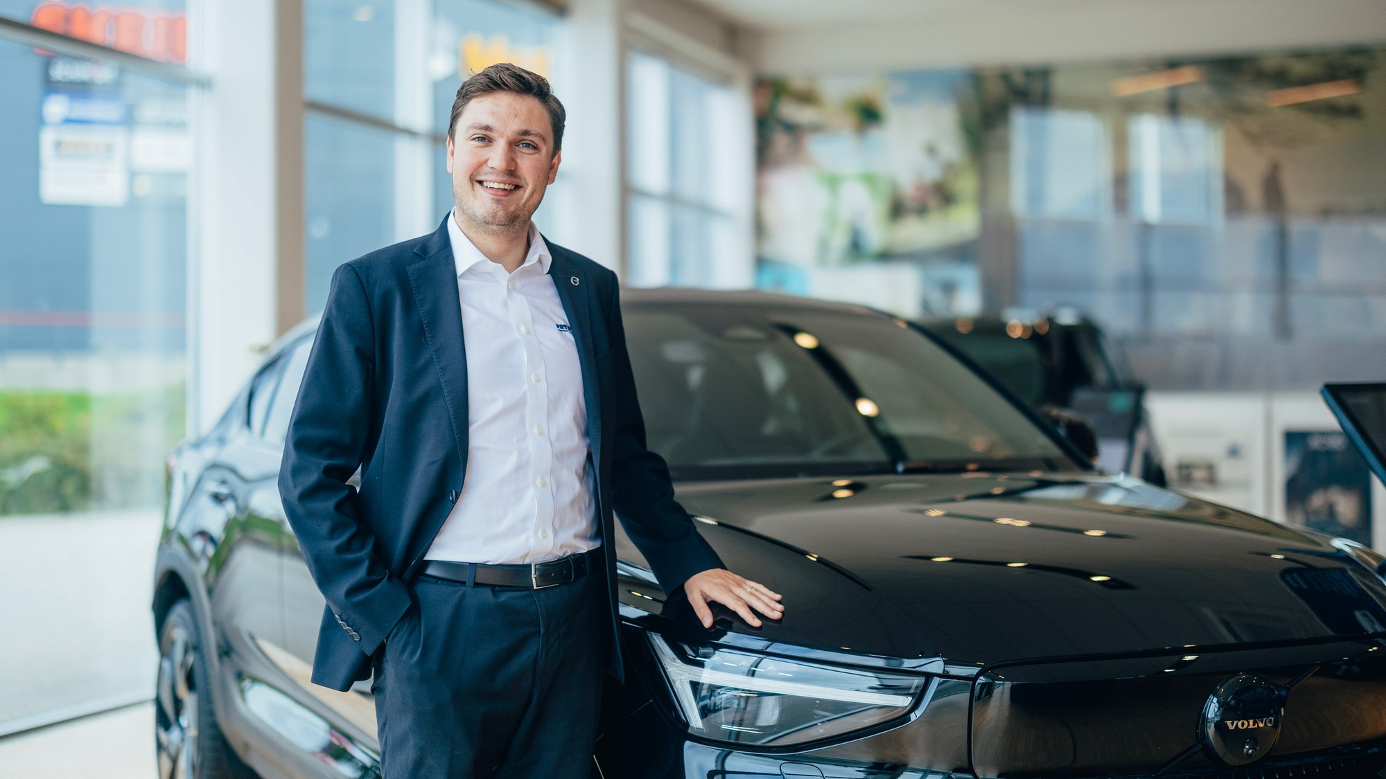 En smilende bilselger i dress står ved siden av en svart Volvo inne i Frydenbø Bils lyse og moderne bilhall i Kristiansand. Han hviler hånden på panseret og ser rett i kamera.
