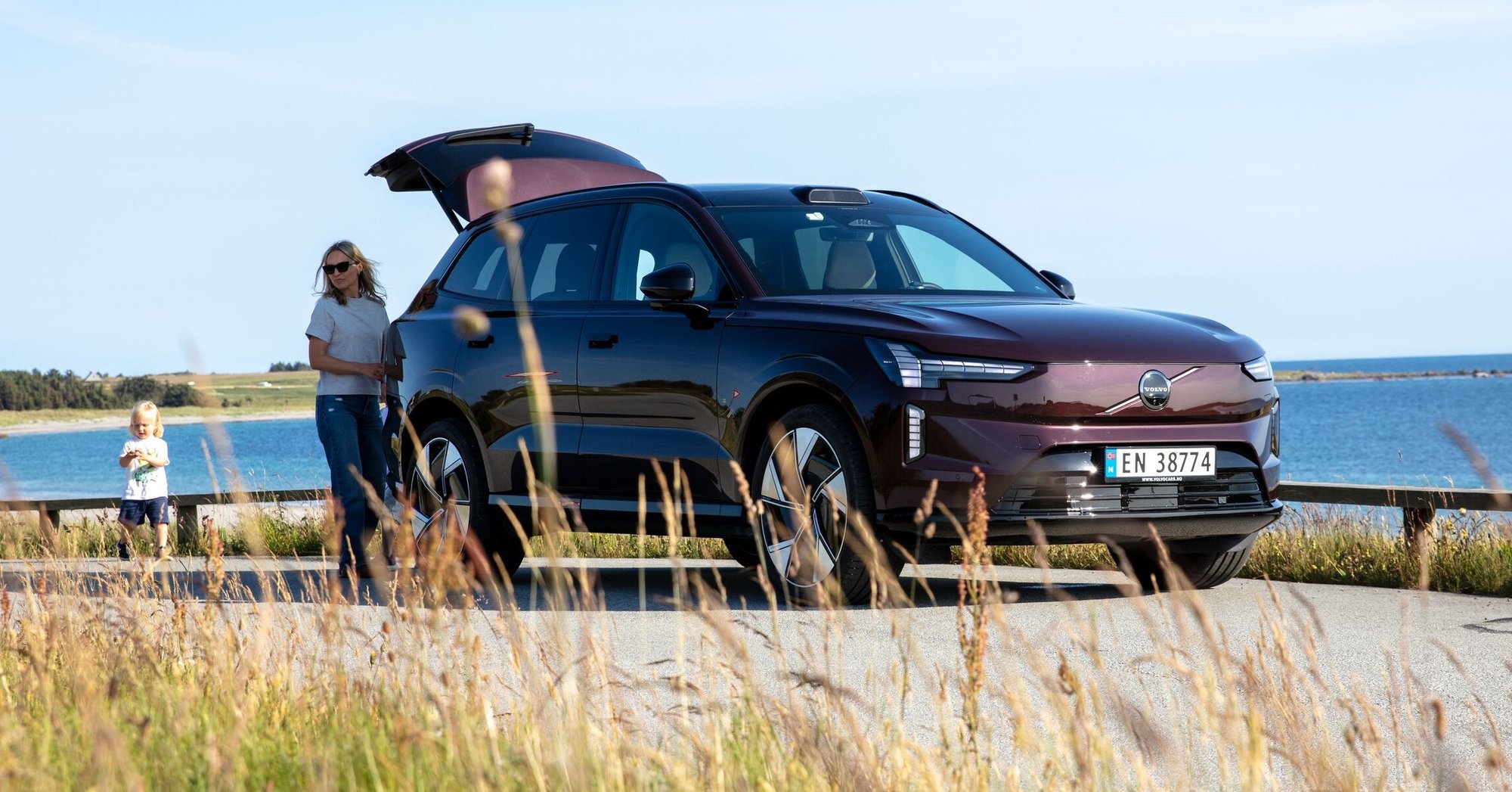 En Mulberry Red Volvo EX90 sett skrått forfra med en dame ved bagasjerommet sammen med et barn. strå i forkant og havet i bakgrunnen