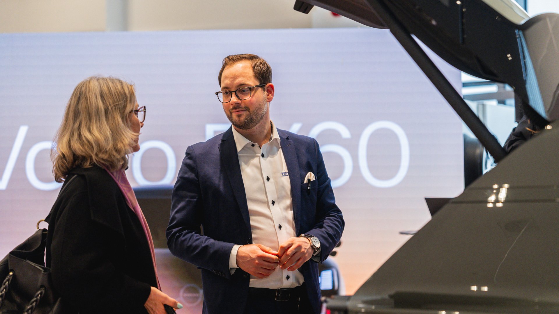 En Frydenbø Bil ansatt snakker med kunde med bagasjerommet til Volvo EX60 i forgrunnen, og en videovegg i bakgrunnen med Volvo EX60 tekst