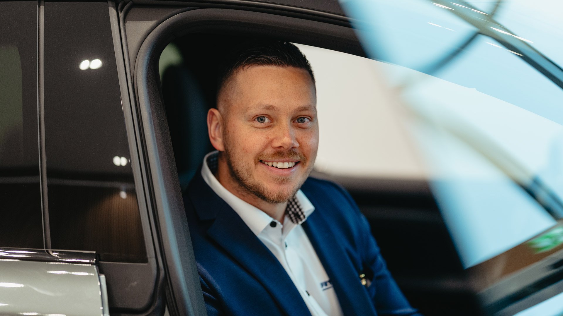 En smilende bilselger sitter i en bil og ser i kamera hos Frydenbø Bil Kokstad
