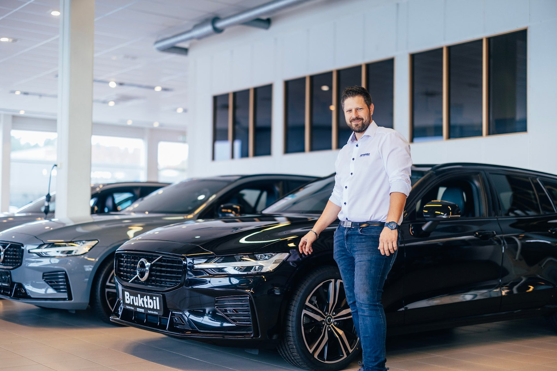 En smilende bilselger hos Frydenbø Bil Kristiansand står i bruktbilhallen siden av en Volvo V60