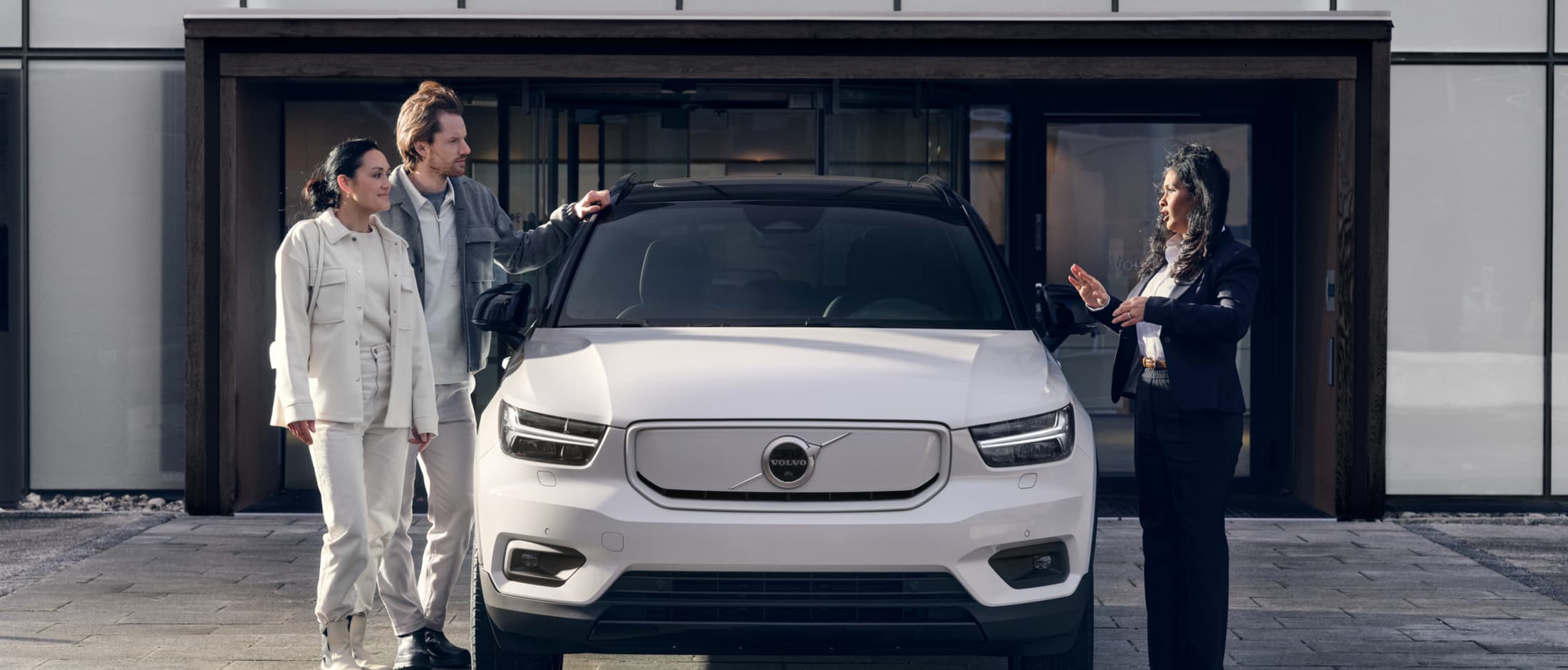 Selger står med kunder og leverer ut deres nye Volvo til dem. Hvit XC40 sett forfra. 
