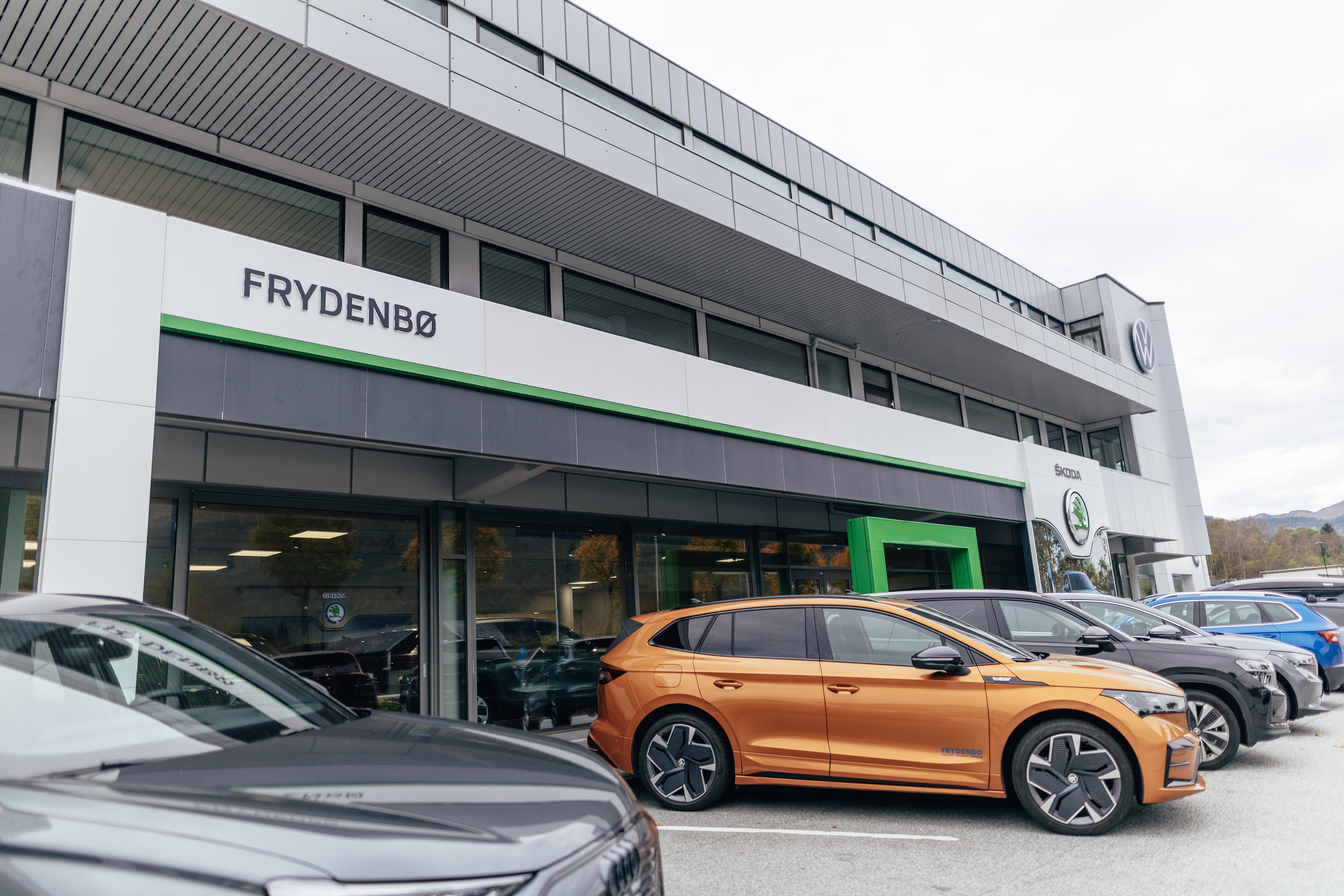 Orange Skoda Enyaq parkert utenfor Frydenbø Bil sin Skoda avdeling i Førde. Store glassruter og innbydende inngang. 