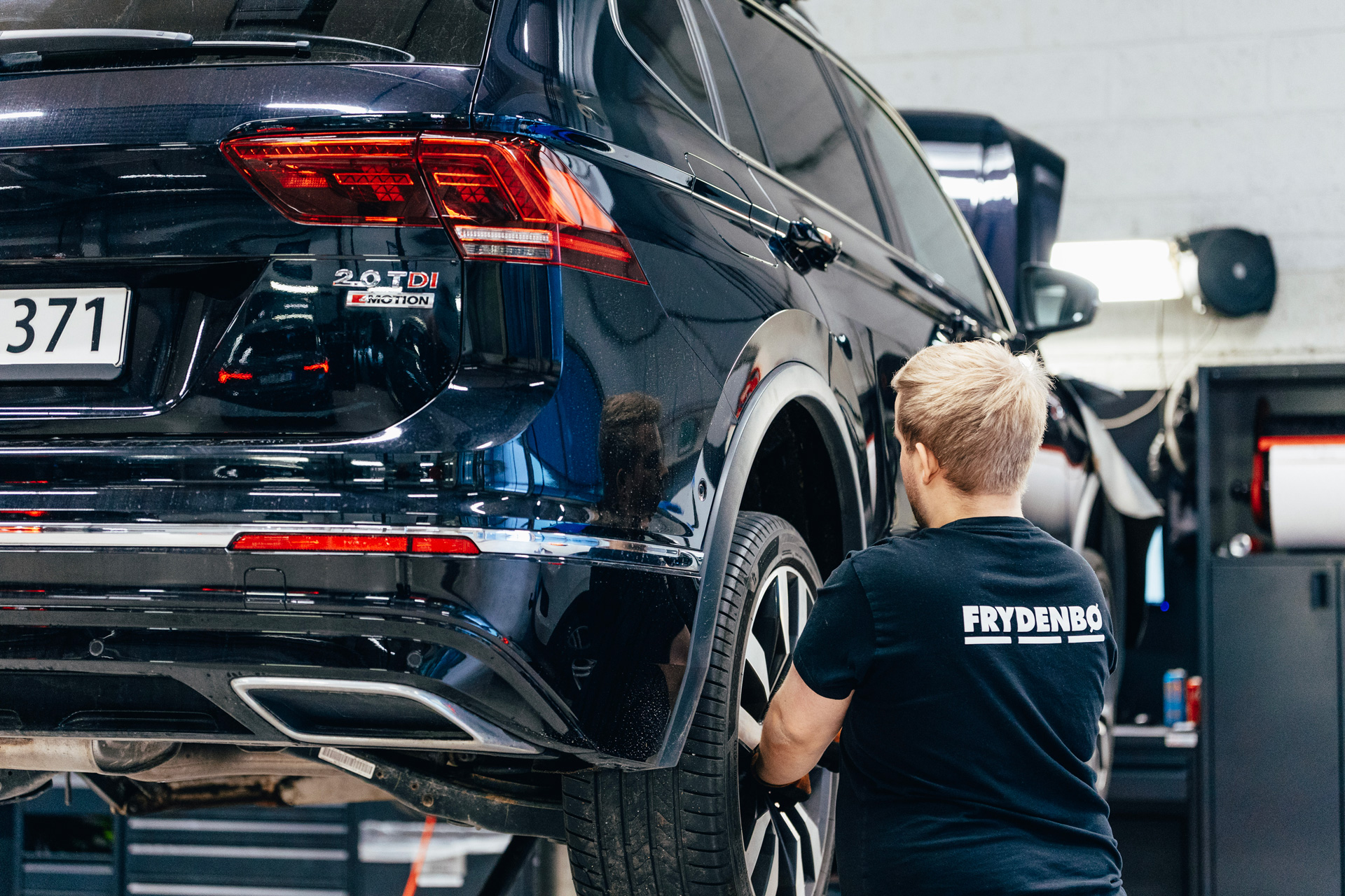 En Volkswagen sett bakfra står på en bukk på verkstedet med en Frydenbø Bil mekaniker som jobber bak på ene hjulet