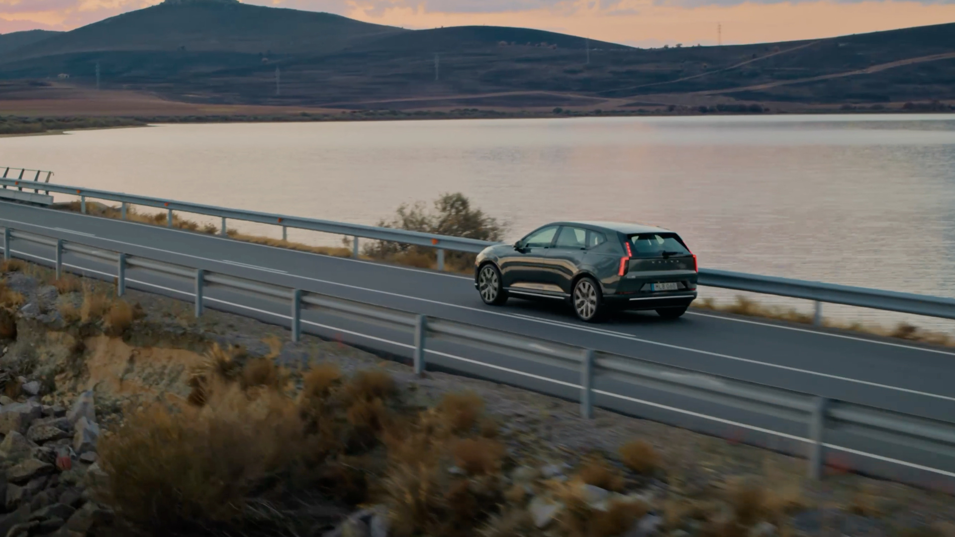 En Volvo EX60 kjører langs et vann på en landevei, sett skrått bakfra