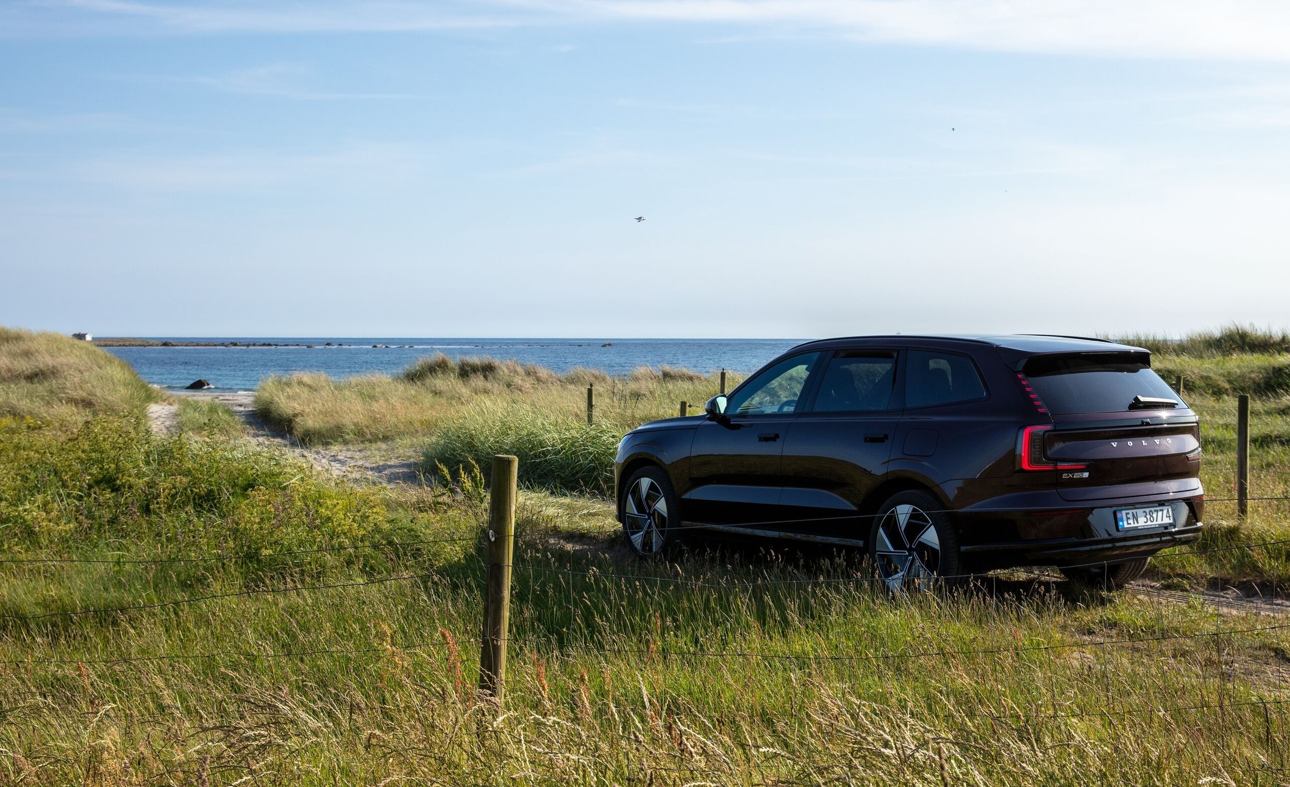 En Mulberry red Volvo EX90 står parkert i gresset med en strand og havet i bakgrunnen