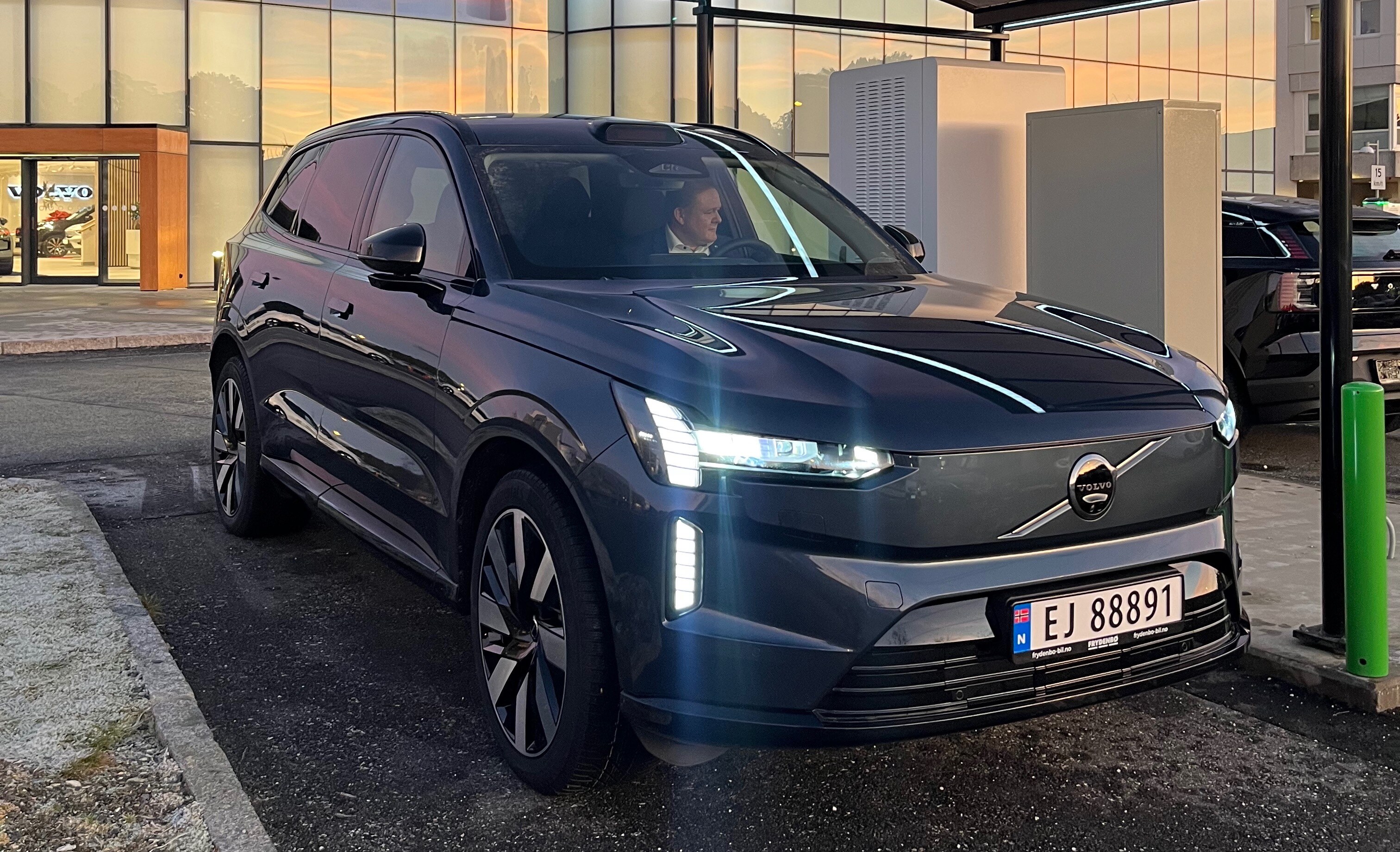 Volvo EX90 står parkert utenfor Frydenbø Bil sitt hovedbygg på Kokstad, Bergen. En av selgerne sitter inni bilen og smiler. 