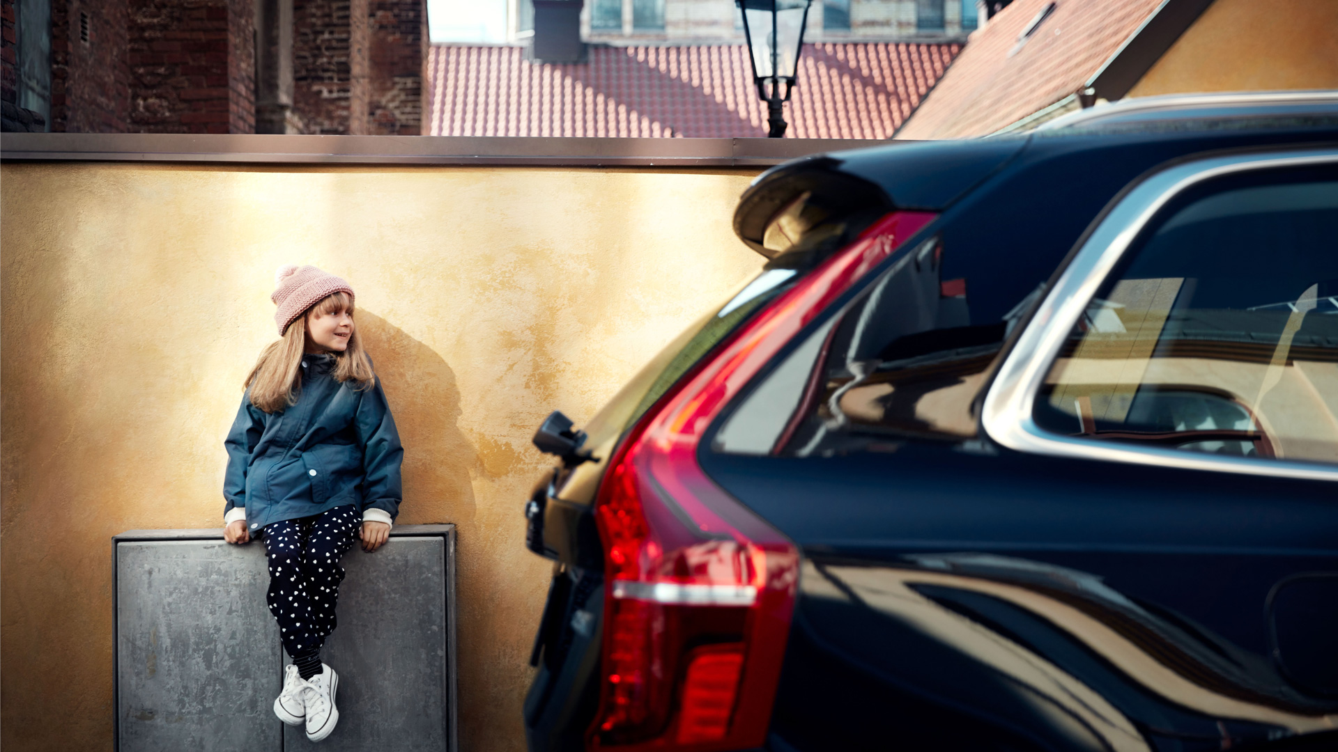 En jente sitter på en strømboks på et fortau og smiler med en bil forgrunnen