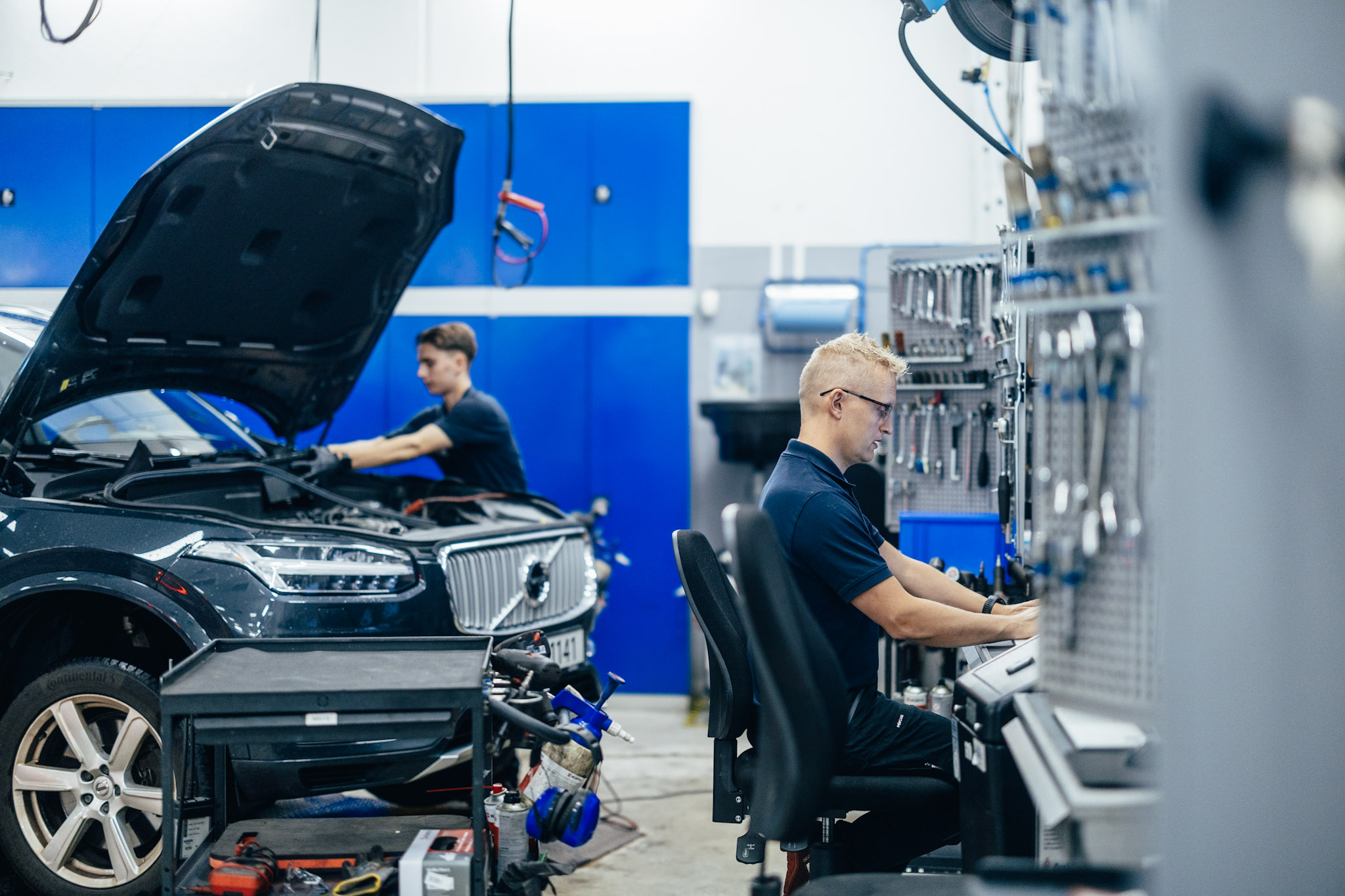 En personlig servicetekniker jobber på en datamaskin mens en annen jobber under panseret på en Volvo XC90 i bakgrunnen