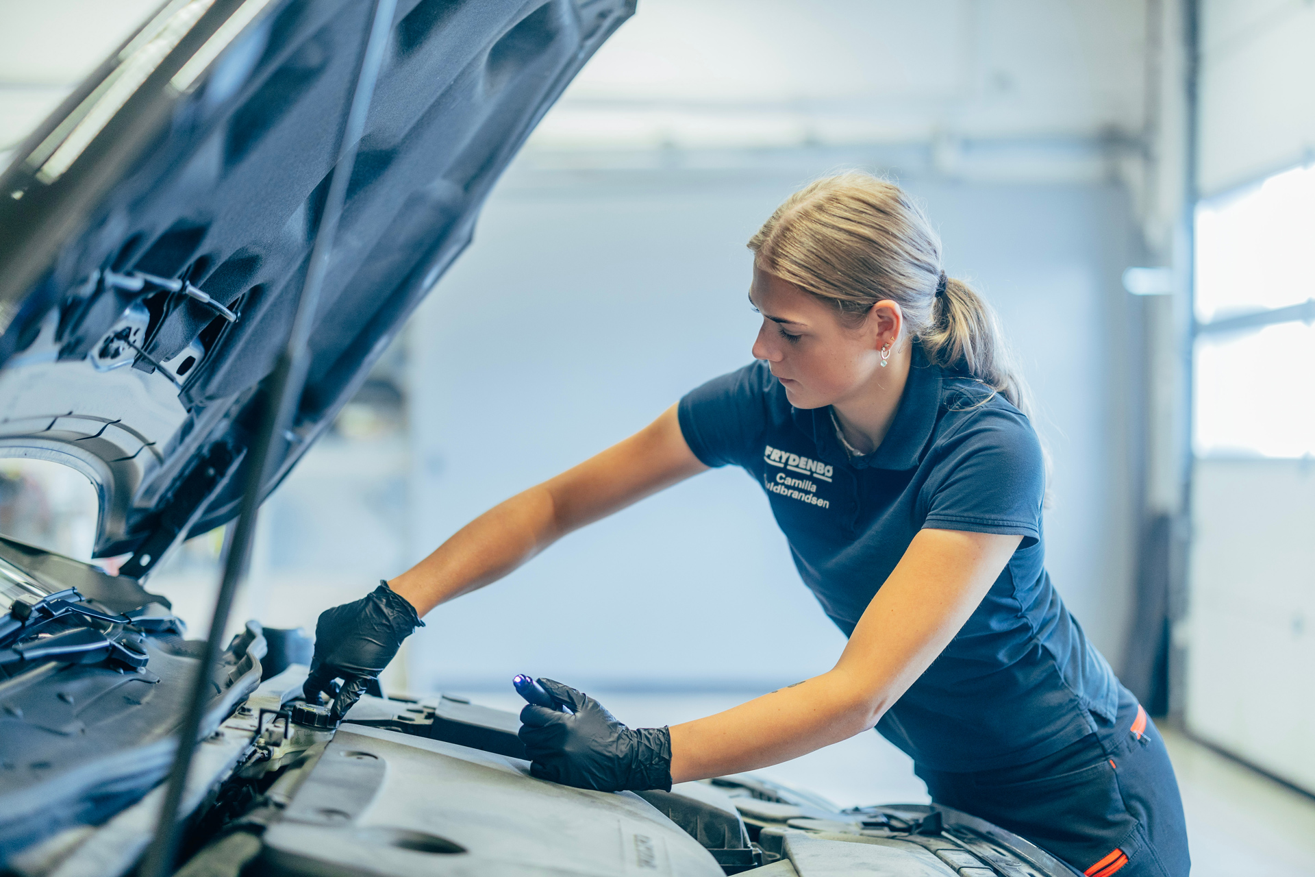 Kvinnelig mekaniker jobber under panseret på en Volvo på et Frydenbø Bil verksted