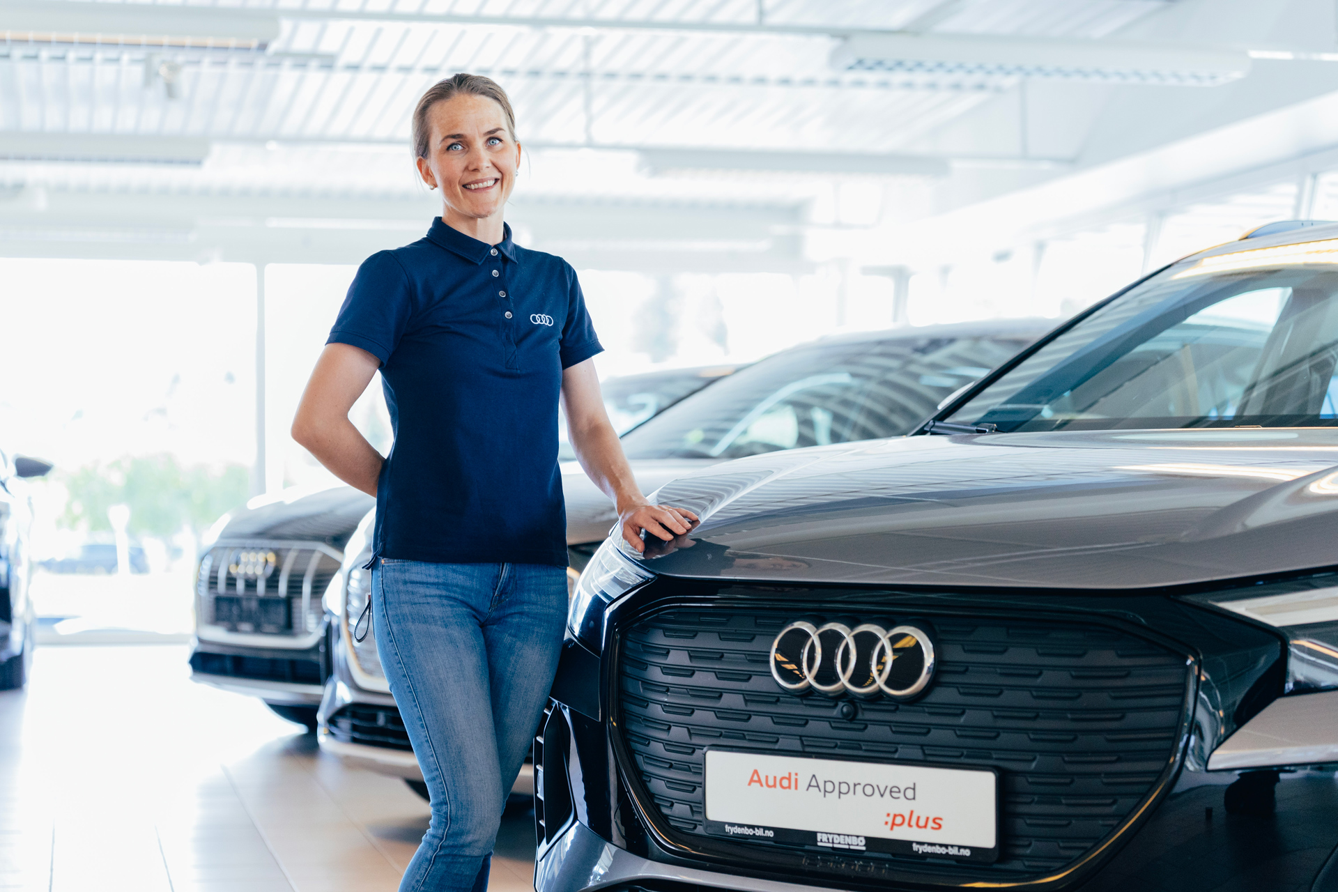 En smilende kvinnelig bilselger hos Frydenbø Bil som står sammen med en Audi Approved :plus bruktbil